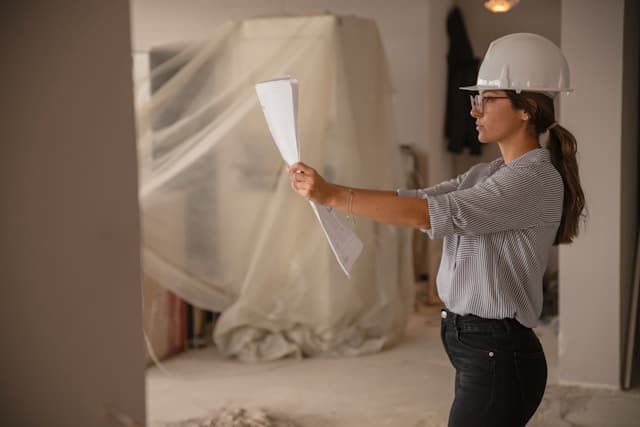 Iemand met een bouwhelm houdt een plan omhoog in een gestripte woning.