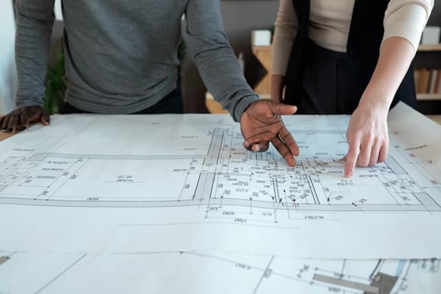 Twee mensen wijzen naar een bouwplan met maatvoering op tafel.
